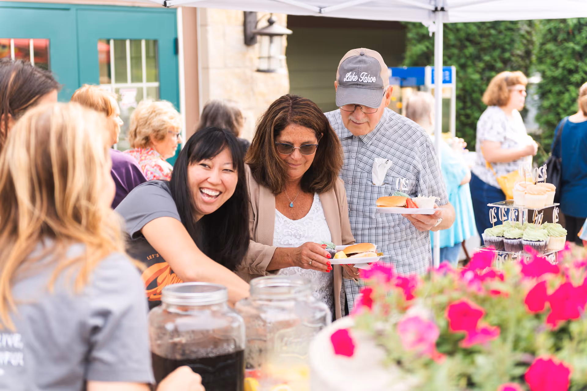 A group of people enjoying an outdoor social event with food and drinks. A woman with long black hair is smiling and serving beverages from large glass containers. Two other people, a woman with sunglasses and a man wearing a cap, are holding plates with sandwiches. There are colorful flowers in the foreground and other people mingling in the background near a building entrance.