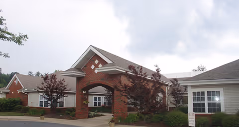 Exterior view of a senior living facility building with a covered entrance supported by brick columns. The building has a combination of brick and siding with multiple windows and a gabled roof. There are small trees and shrubs planted around the entrance area under a cloudy sky.