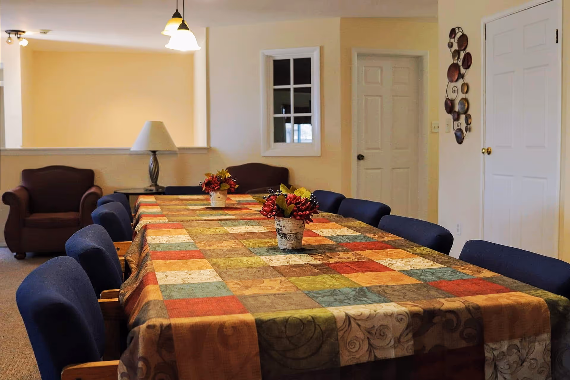 A dining room with a long table covered by a colorful patchwork tablecloth. The table is surrounded by blue cushioned chairs and decorated with two small flower arrangements. In the background, there are two armchairs, a table lamp, a small window, and two closed doors. The walls are painted light yellow and white.