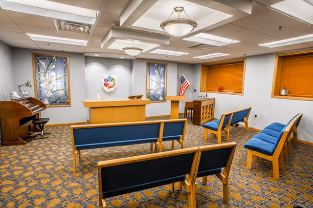 Interior view of a small chapel or meditation room with wooden benches featuring blue cushions, a wooden altar at the front, two stained glass windows on the wall behind the altar, an American flag in the corner, a piano on the left side, and two windows with orange blinds on the right wall.