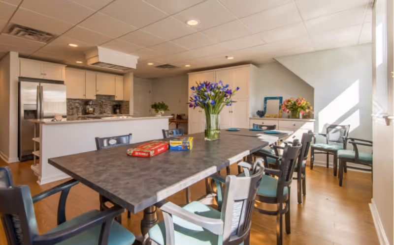 A bright and spacious kitchen and dining area with a large rectangular table surrounded by chairs. The table has a vase with purple flowers and two board games on it. The kitchen features white cabinets, a stainless steel refrigerator, and a countertop with a stove. There is a sideboard with flowers and a mirror against the far wall, and sunlight streams in through a window on the right.