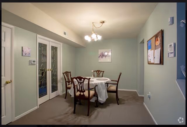 A small dining room with a round table covered with a white tablecloth and four wooden chairs with cushioned seats. The walls are painted light green, and there is a chandelier with multiple lights hanging from the ceiling. On the left side, there are double glass doors with decorative patterns, and on the right wall, there is a bulletin board with papers pinned to it. A thermostat is mounted on the left wall near the doors.
