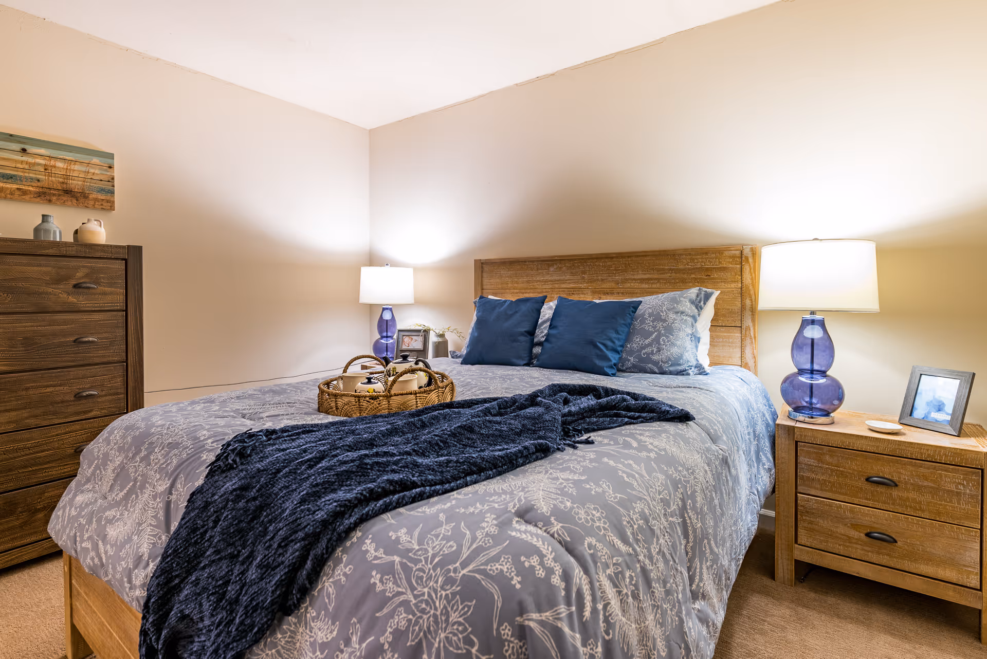 Cozy bedroom with a wooden bed dressed in blue patterned bedding, two nightstands with lamps, and a dresser.