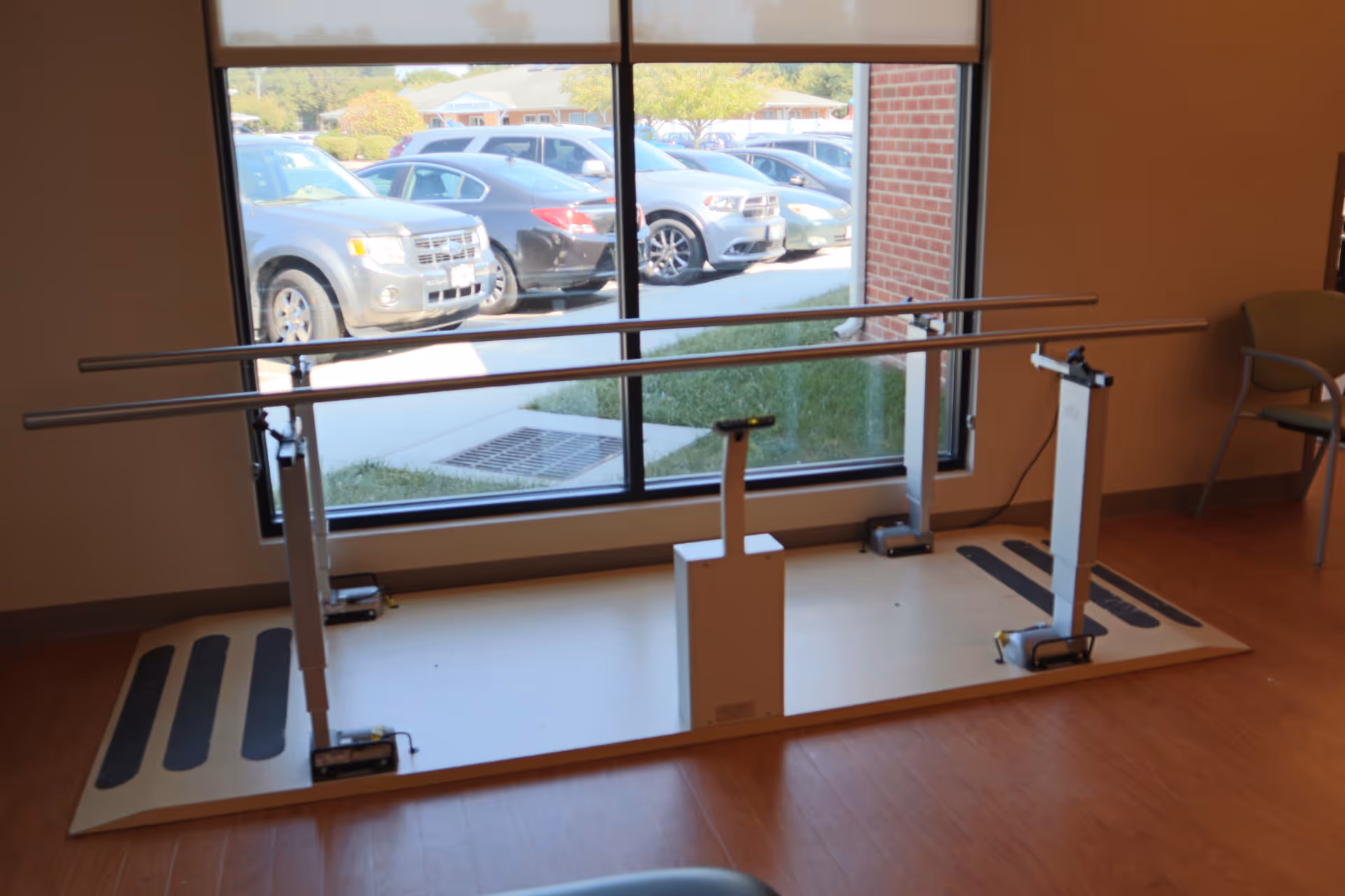 A physical therapy parallel bar setup in a room with large windows showing parked cars outside. The bars are mounted on a platform with non-slip strips, and there is a chair visible to the right.