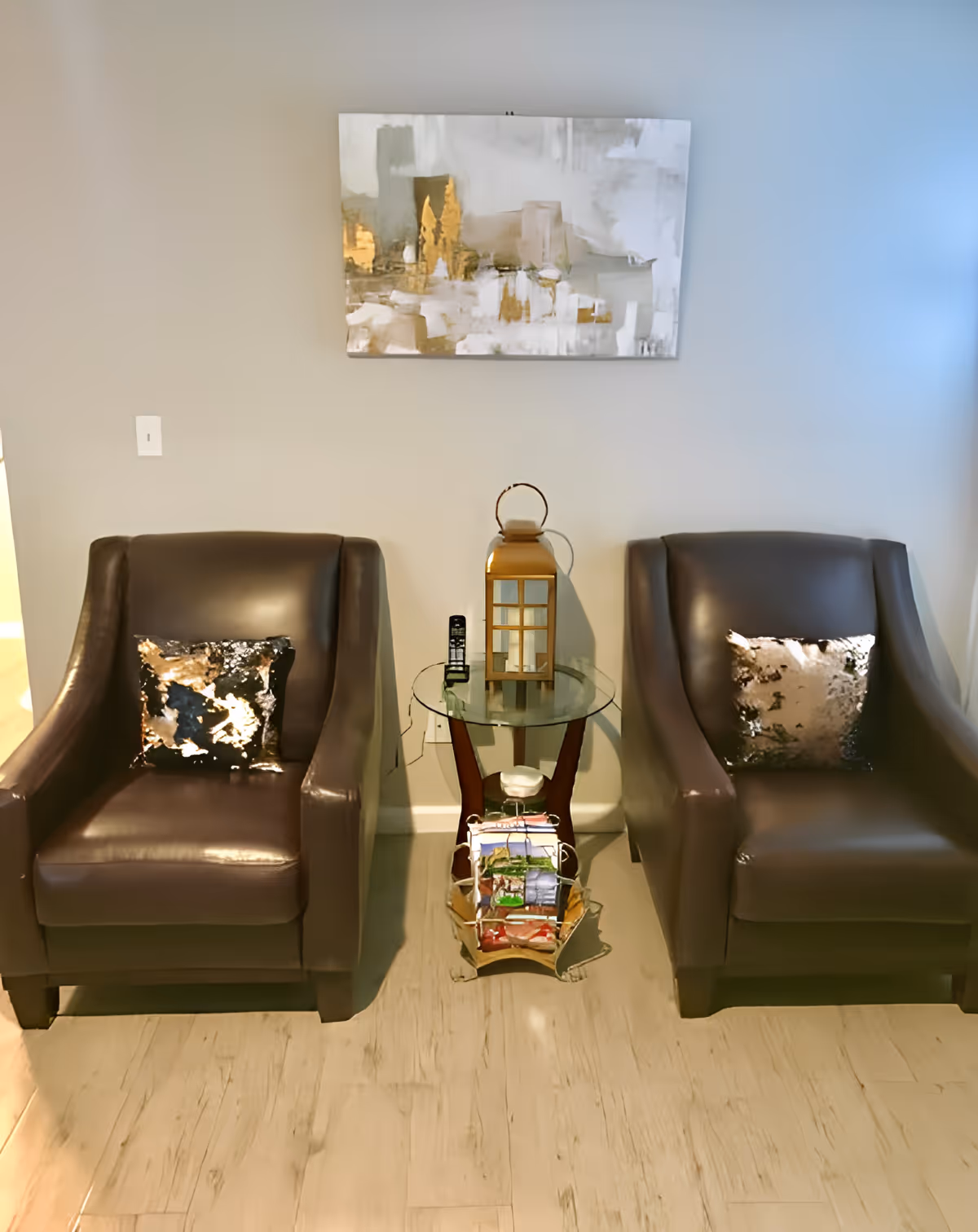 Two dark brown leather armchairs with metallic decorative pillows are placed on either side of a small glass-top side table. On the table is a decorative lantern and a cordless phone. Below the table is a magazine rack with various magazines. A modern abstract painting with gold and white tones hangs on the light gray wall behind the chairs.