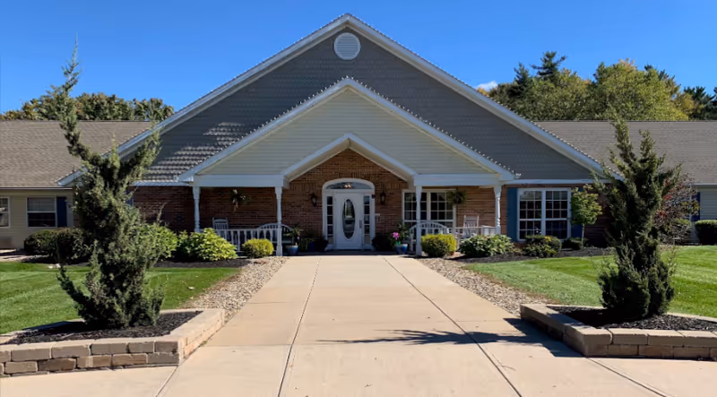 Front exterior view of a single-story brick and siding building with a peaked roof, a covered entrance with white columns, a glass door, and landscaped greenery including bushes and small trees on either side of a wide concrete walkway.