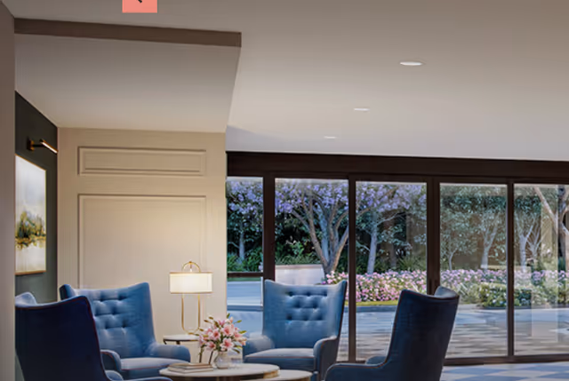 A cozy seating area with four blue upholstered armchairs arranged around a small round table with a flower vase. Behind the seating area, large floor-to-ceiling windows provide a view of a garden with trees and flowering plants.