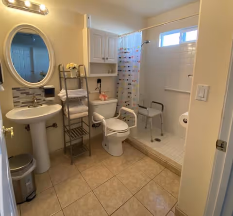 A clean and accessible bathroom with beige tiled floor, a white pedestal sink with an oval mirror above it, a metal rack holding folded towels and a small flower vase, a toilet with safety rails, and a walk-in shower with a white tiled floor, grab bars, a shower chair, and a colorful polka dot shower curtain. The walls are light-colored and there is a small window in the shower area.
