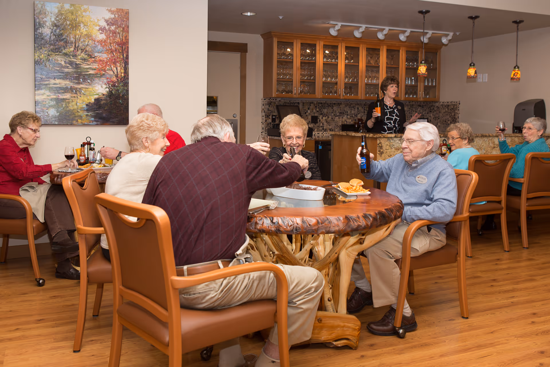 A group of elderly people sitting around a wooden table in a dining area, enjoying drinks and socializing. In the background, a woman stands behind a counter with glass cabinets filled with glassware. The room has warm lighting and wooden flooring, with a painting of a nature scene on the wall.