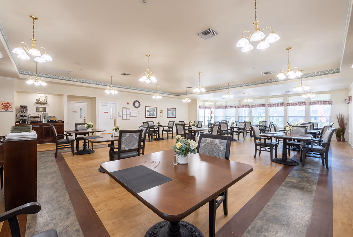 A spacious dining room with multiple wooden tables and chairs arranged neatly. Each table has a small flower arrangement. The room is well-lit with several ceiling light fixtures and large windows along one wall letting in natural light. There is a coffee and beverage station against one wall and framed pictures and a clock on the walls.
