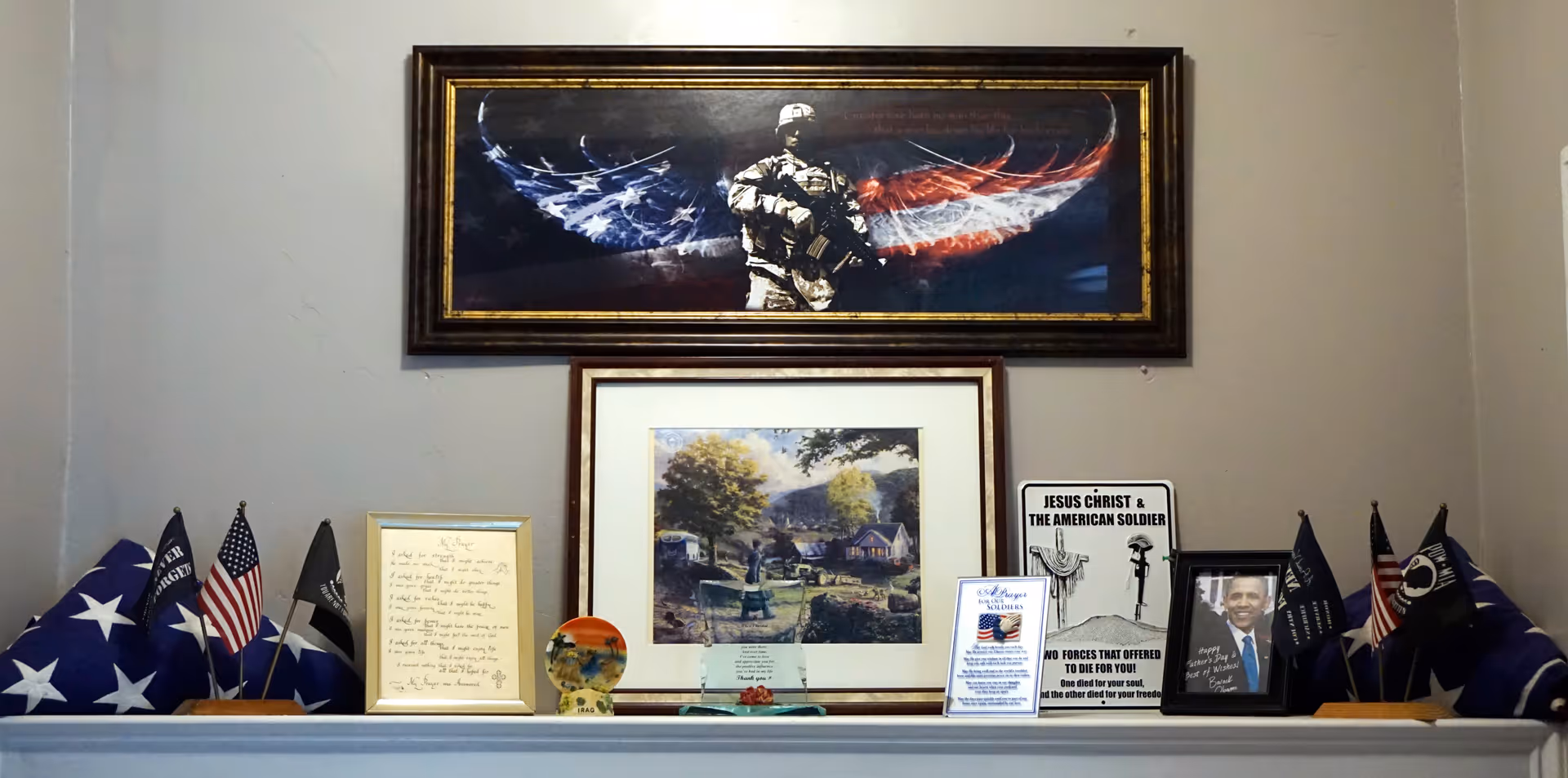 A display shelf featuring patriotic and military-themed items including folded American flags, small American and POW/MIA flags, framed pictures, plaques, and a sign that reads 'Jesus Christ & The American Soldier'. Above the shelf are two framed artworks: one of a soldier with wings in the colors of the American flag, and another depicting a peaceful countryside scene.