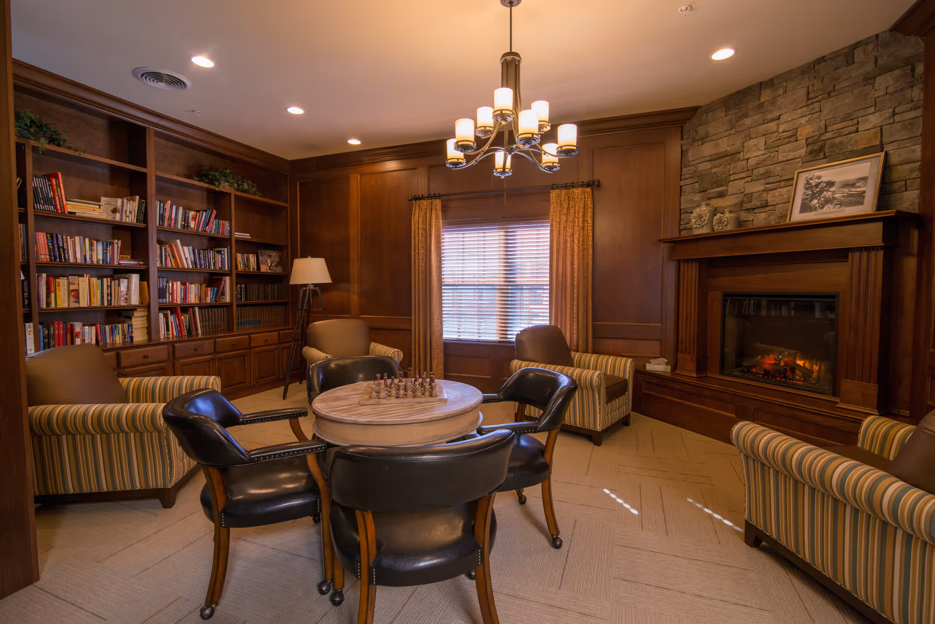 A cozy common room with a round table and four black leather chairs in the center, a chessboard on the table, four striped armchairs around the room, a stone fireplace with a wooden mantle on the right, a large window with curtains in the center, and a wooden bookshelf filled with books on the left. The room has warm wood paneling and a chandelier hanging from the ceiling.