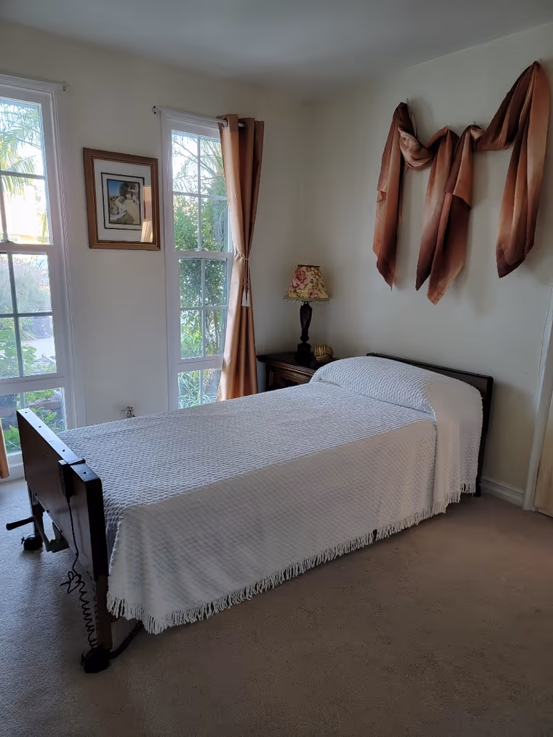 A single adjustable bed with a white bedspread in a sunlit bedroom with tall windows, a nightstand and wall drapery.
