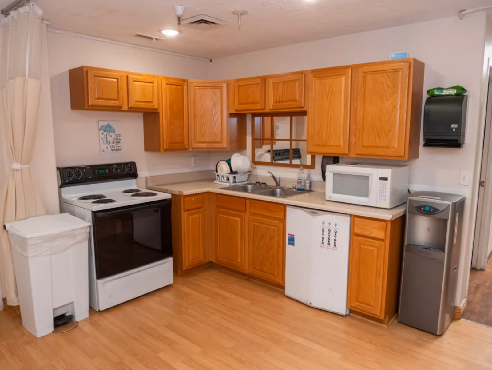 Small kitchen area with oak cabinets, an electric stove, sink with dish rack, microwave, mini-fridge, water cooler and trash can.