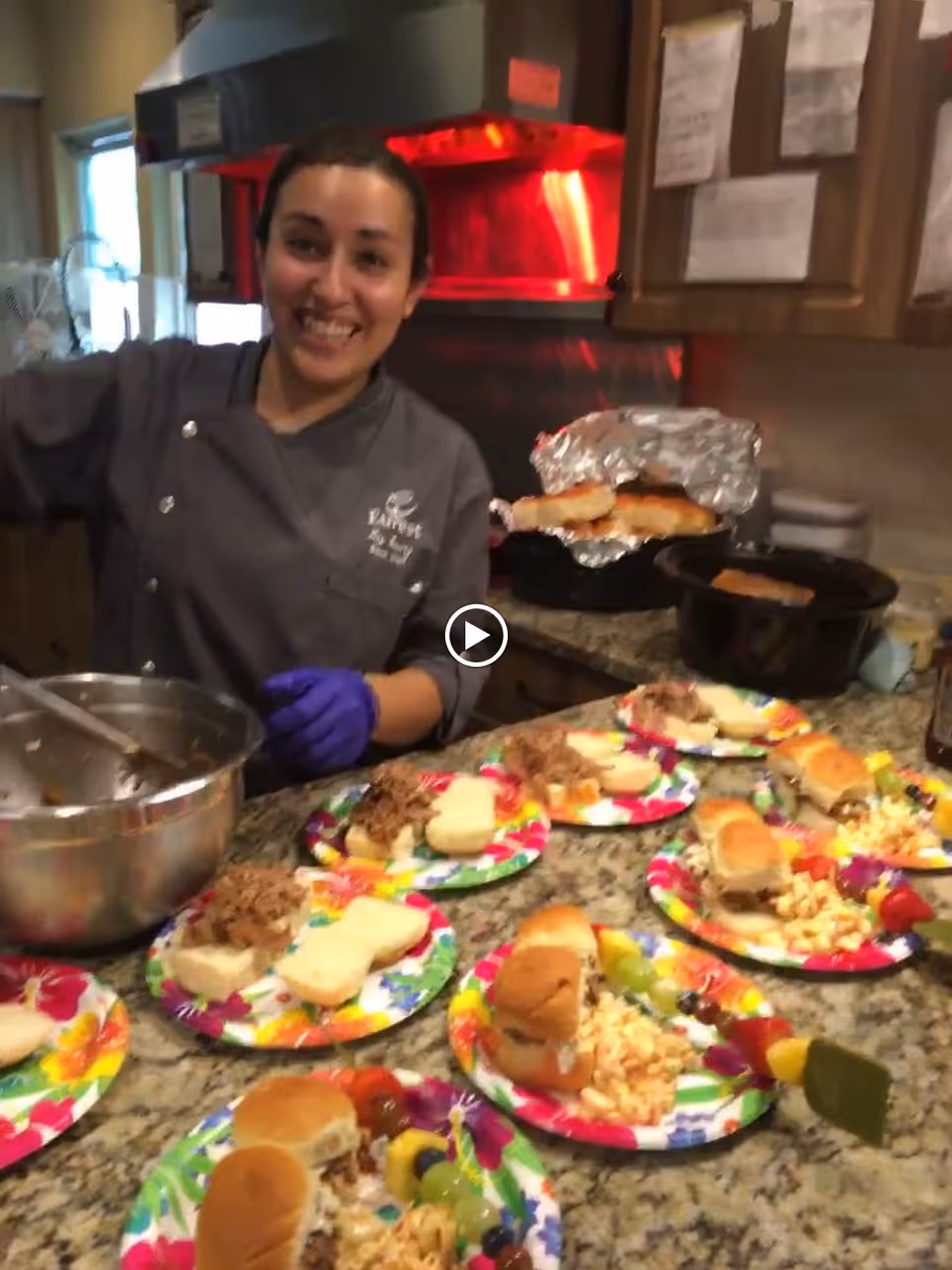 A smiling woman wearing a gray chef coat and purple gloves stands behind a kitchen counter with several colorful plates of food, including sandwiches and fruit skewers. The kitchen has wooden cabinets and a red-lit oven in the background.