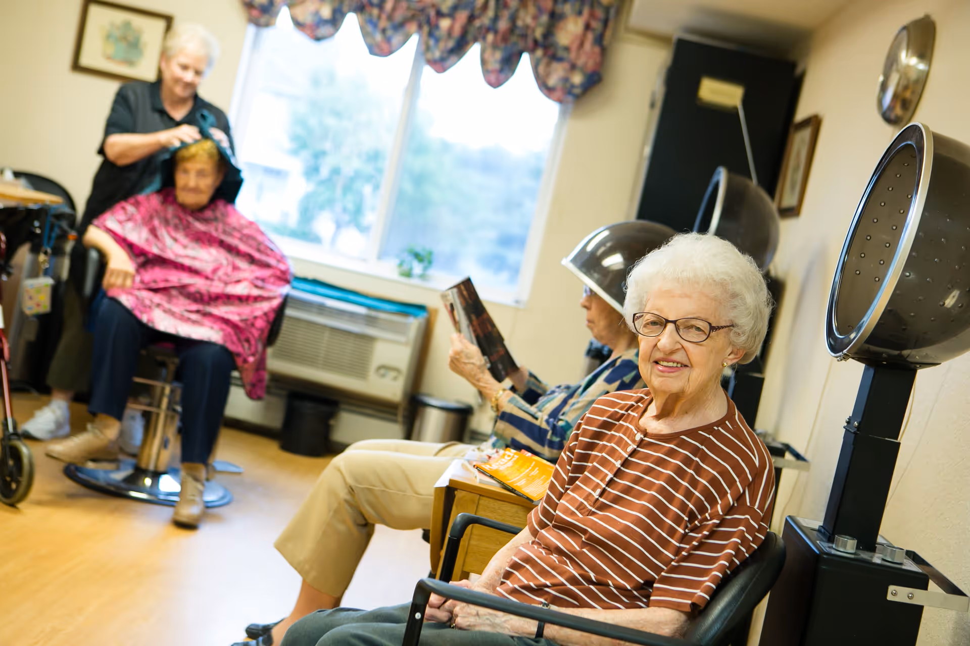 An elderly woman in a striped shirt smiling at the camera while sitting in a chair under a hair dryer in a salon area. In the background, another elderly woman is getting her hair done by a stylist, and a third woman is sitting under a hair dryer reading a magazine. The room has a large window with floral curtains and light-colored walls.