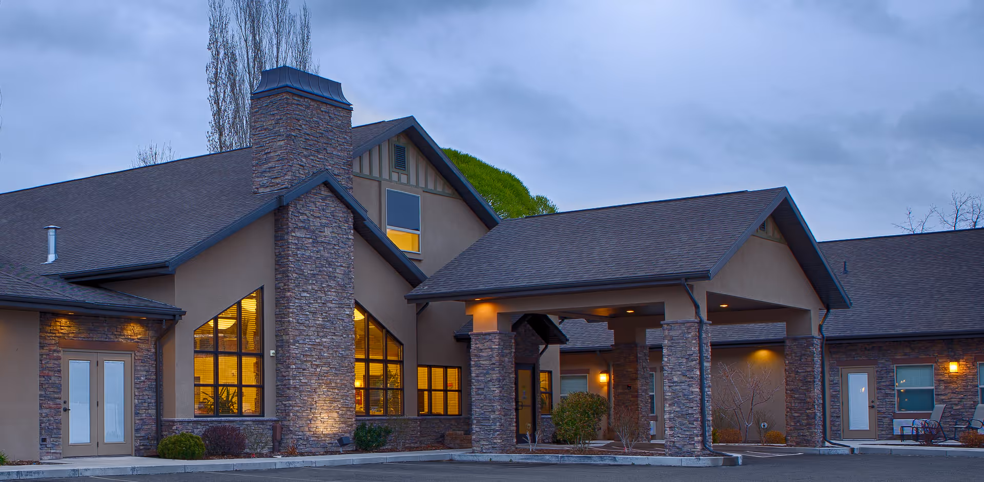 Exterior view of Pheasant View Assisted Living facility during dusk, showing a building with stone and beige stucco walls, large windows with warm interior lighting, a covered entrance supported by stone pillars, and a paved parking area in front.
