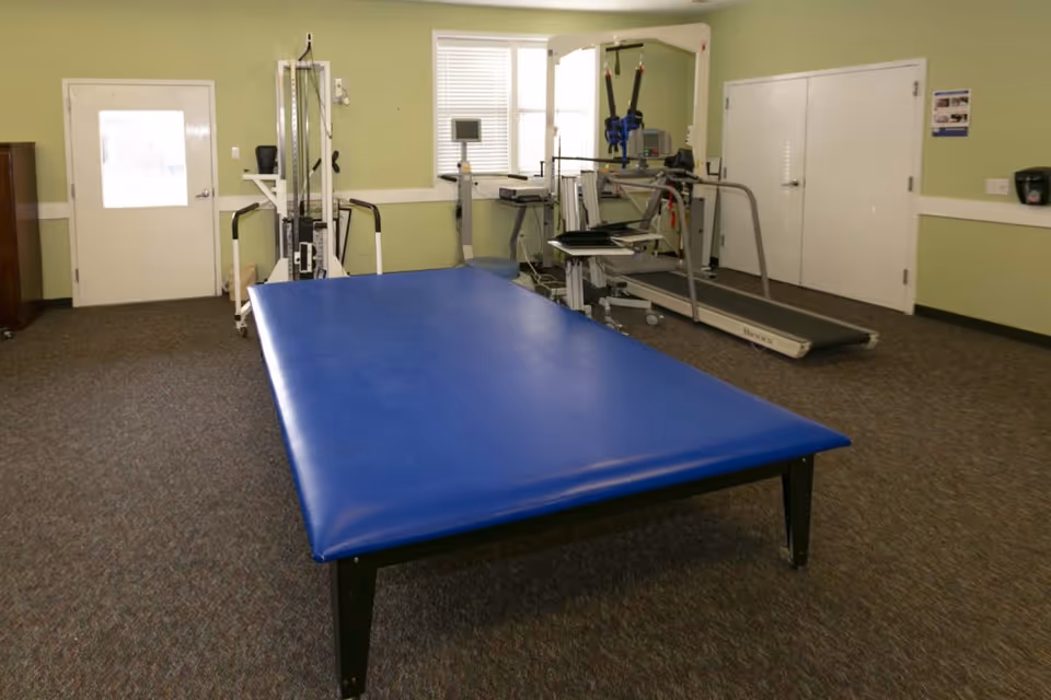 Physical therapy room with a large blue treatment table in the foreground and exercise equipment including a treadmill in the background.