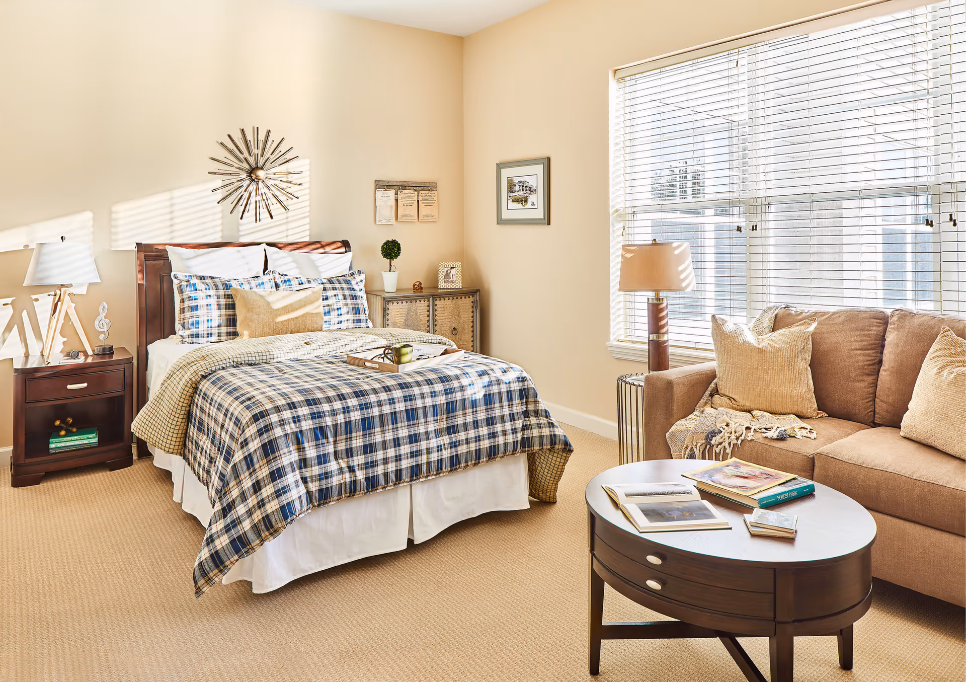 A bright bedroom with a bed covered in blue and white plaid bedding, a wooden nightstand with a lamp and decorative items, a small cabinet with framed pictures and a plant, a brown sofa with beige pillows, a round wooden coffee table with books, and a large window with white blinds letting in natural light.