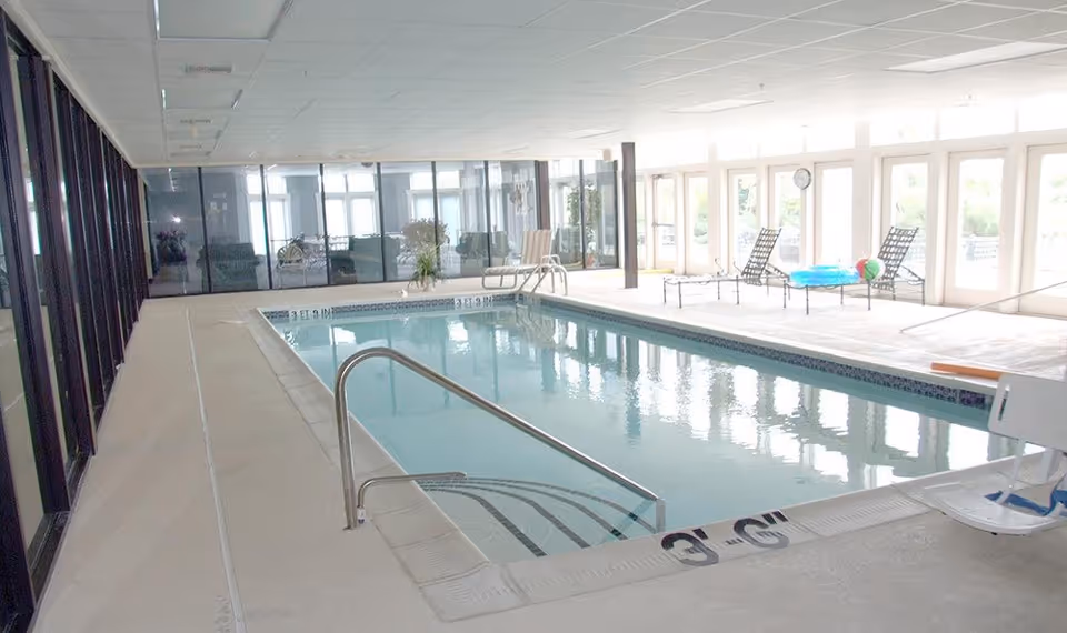 Indoor swimming pool with clear water, metal handrails, and steps leading into the pool. The pool area is surrounded by large windows letting in natural light, with lounge chairs and pool toys on the far side. The ceiling has recessed lighting and the floor is tiled.