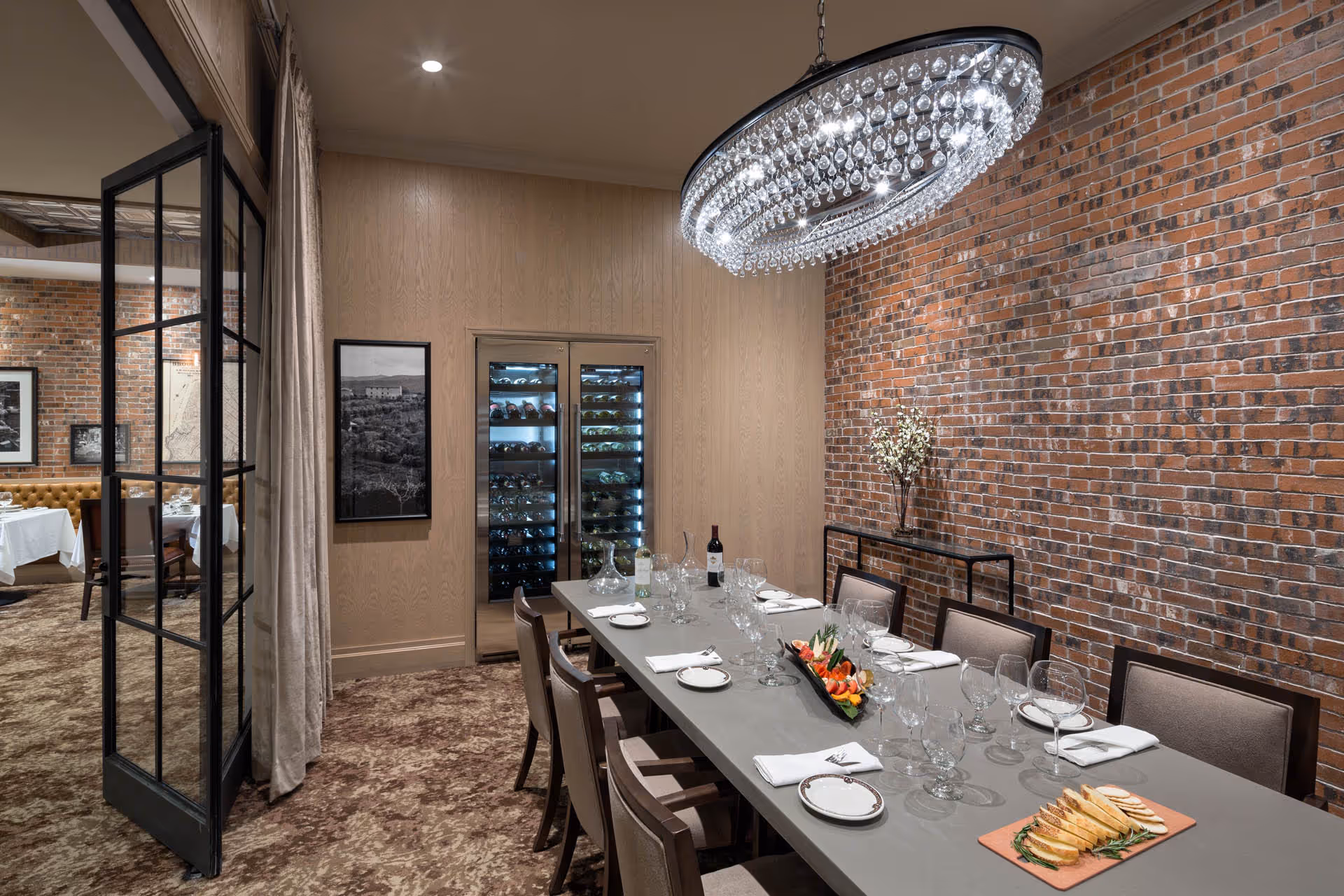 A private dining room with a long rectangular table set for a meal, featuring plates, wine glasses, napkins, and a centerpiece of sliced bread and vegetables. The room has a brick accent wall, a modern chandelier with hanging crystals, a wine cooler, and framed black and white photos on the walls. A glass door opens to an adjacent dining area with more tables and chairs.