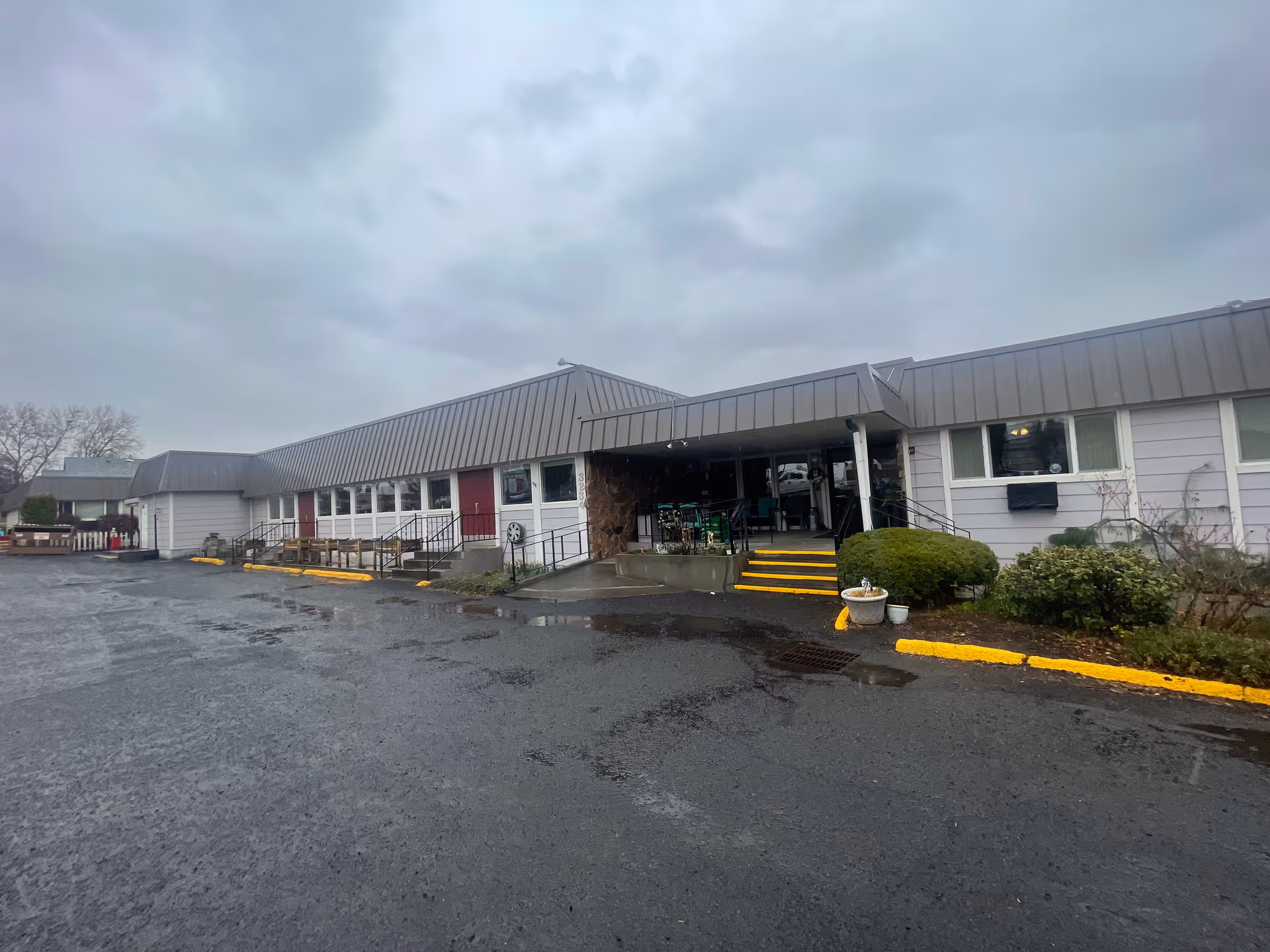Exterior view of a single-story building with a gray metal roof and light purple siding under a cloudy sky. The building has several windows and red doors, with steps and a ramp leading to a covered entrance area. There are some bushes and planters near the entrance, and a wet asphalt parking lot in front.