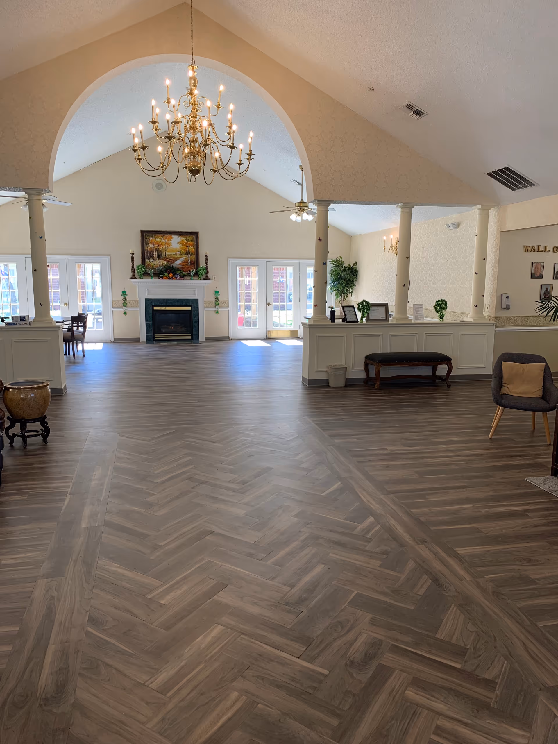 Spacious interior common area with a high vaulted ceiling, a large chandelier, and a fireplace centered on the far wall. The room features wood-patterned flooring, white pillars, and multiple glass doors letting in natural light. There are some chairs, a bench, plants, and framed pictures on the walls.