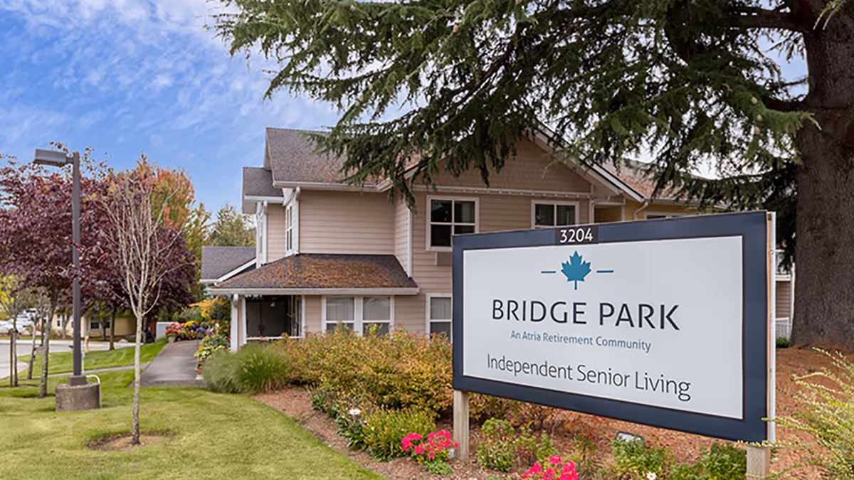 Exterior view of a senior living facility named Bridge Park, showing a large sign with the facility's name and description 'Independent Senior Living' in front of a two-story building surrounded by trees and landscaped greenery.