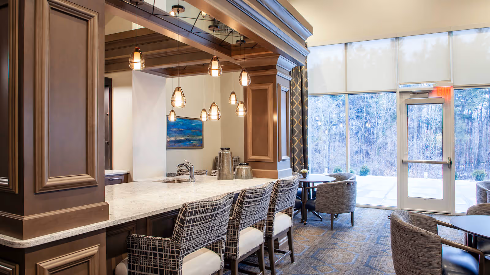 Bright communal lounge and dining area with a marble-topped bar, pendant lights, barstools and seating by large windows overlooking trees.