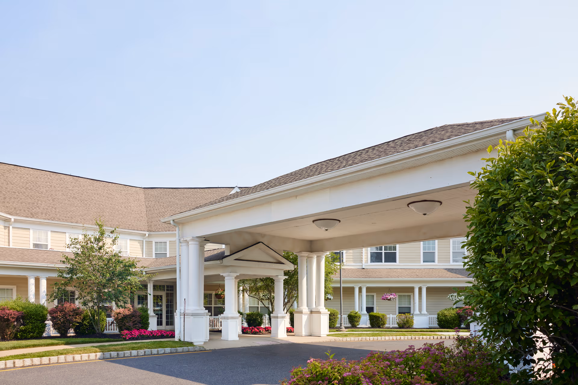 Covered porte-cochere entrance with white columns at the front of a multi-story senior living facility surrounded by landscaping.