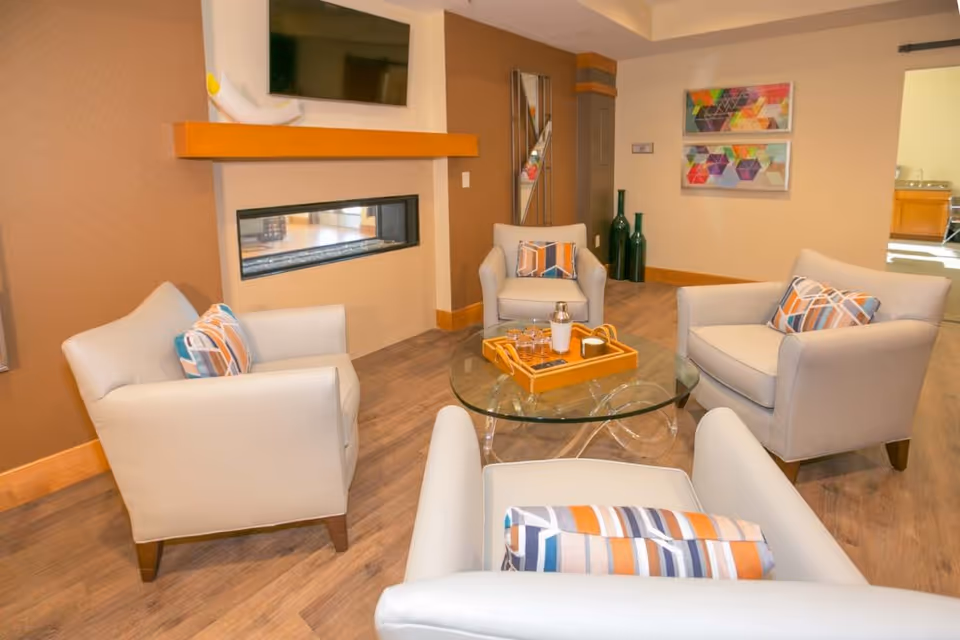 A cozy living room area with four light gray armchairs arranged around a round glass coffee table. The table has a wooden tray with a shaker and glasses. The room features a modern built-in fireplace beneath a mounted flat-screen TV, colorful geometric artwork on the wall, and decorative tall green vases in the corner. The flooring is wood, and the walls are painted in warm neutral tones.