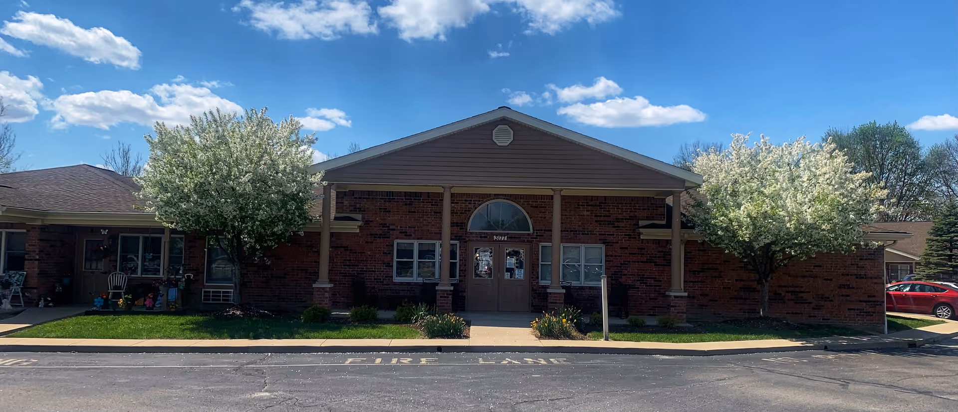Front exterior view of a single-story brick building with a covered entrance supported by columns. Two flowering trees flank the entrance, and a red car is parked to the right. The sky is blue with scattered clouds.