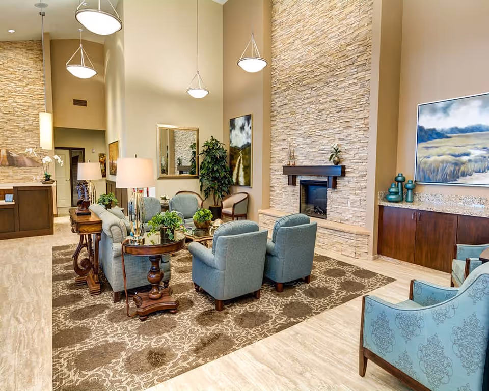 Bright, upscale common lounge with upholstered chairs around a coffee table, a stone fireplace, and tall pendant lights.