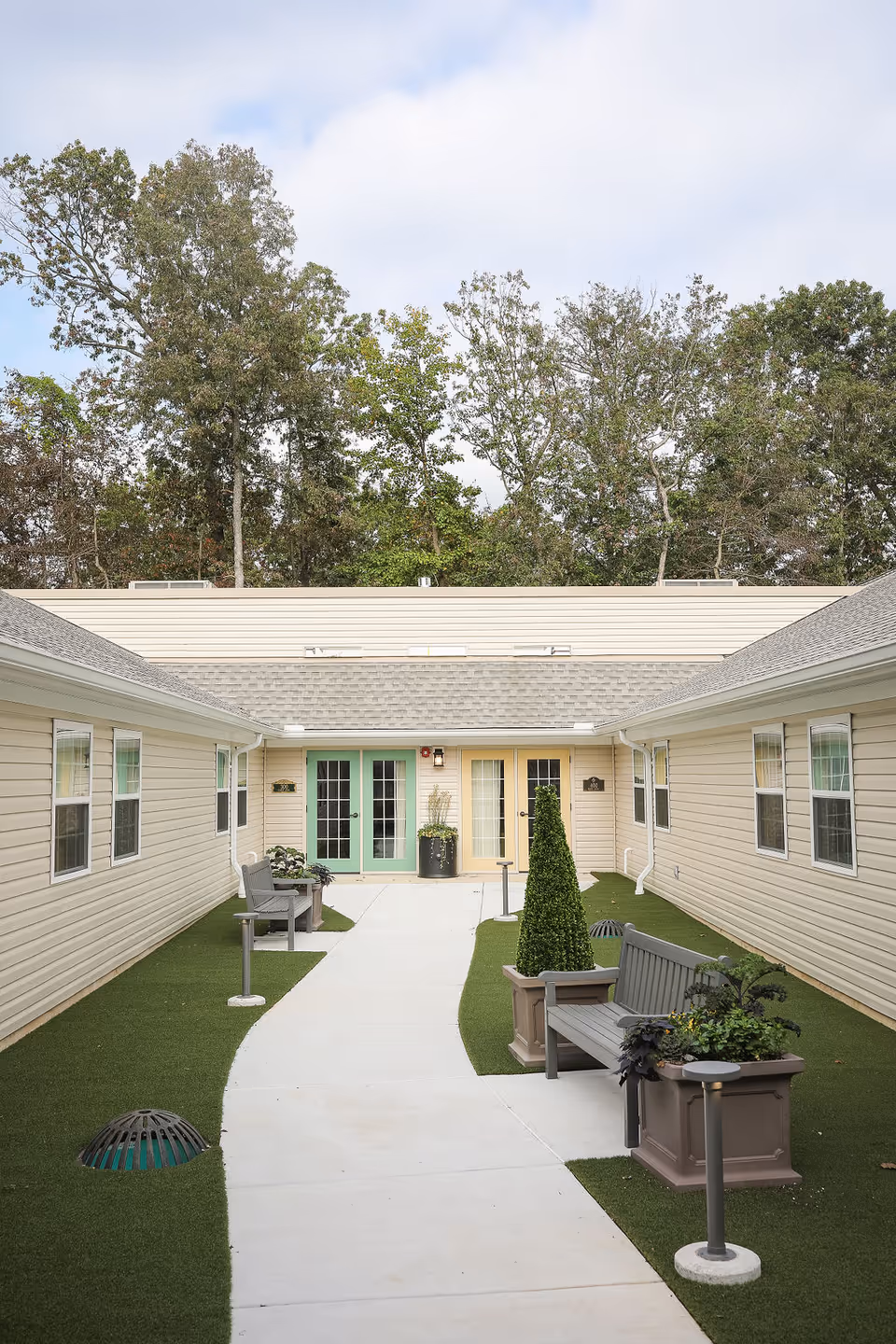 Outdoor courtyard area at Artis Senior Living of Potomac featuring a paved walkway, green artificial grass, benches, potted plants, and beige buildings with multiple windows and doors. Trees are visible in the background under a partly cloudy sky.