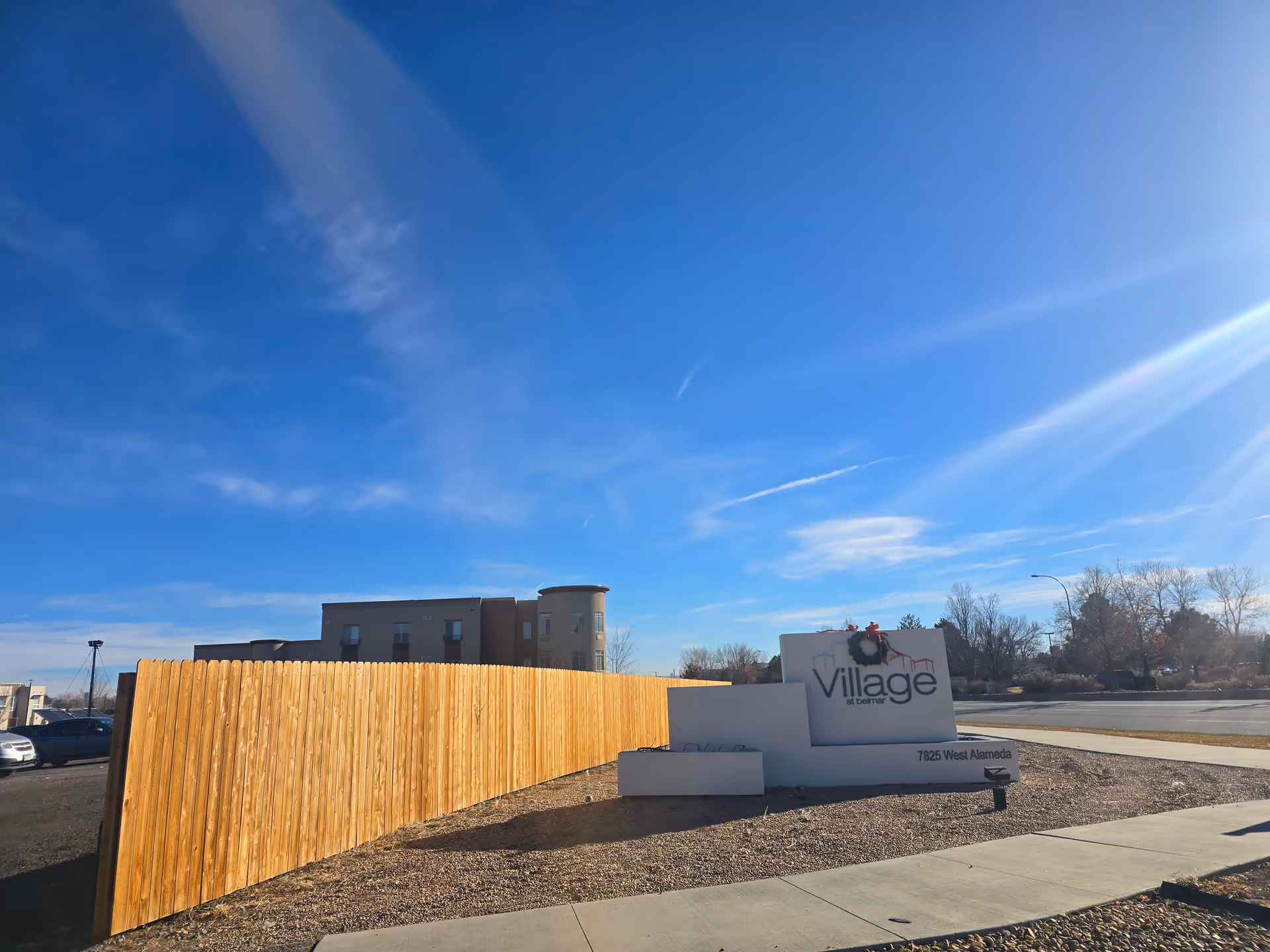 Outdoor view of the Village at Belmar facility sign with a wooden fence and a building in the background under a clear blue sky.
