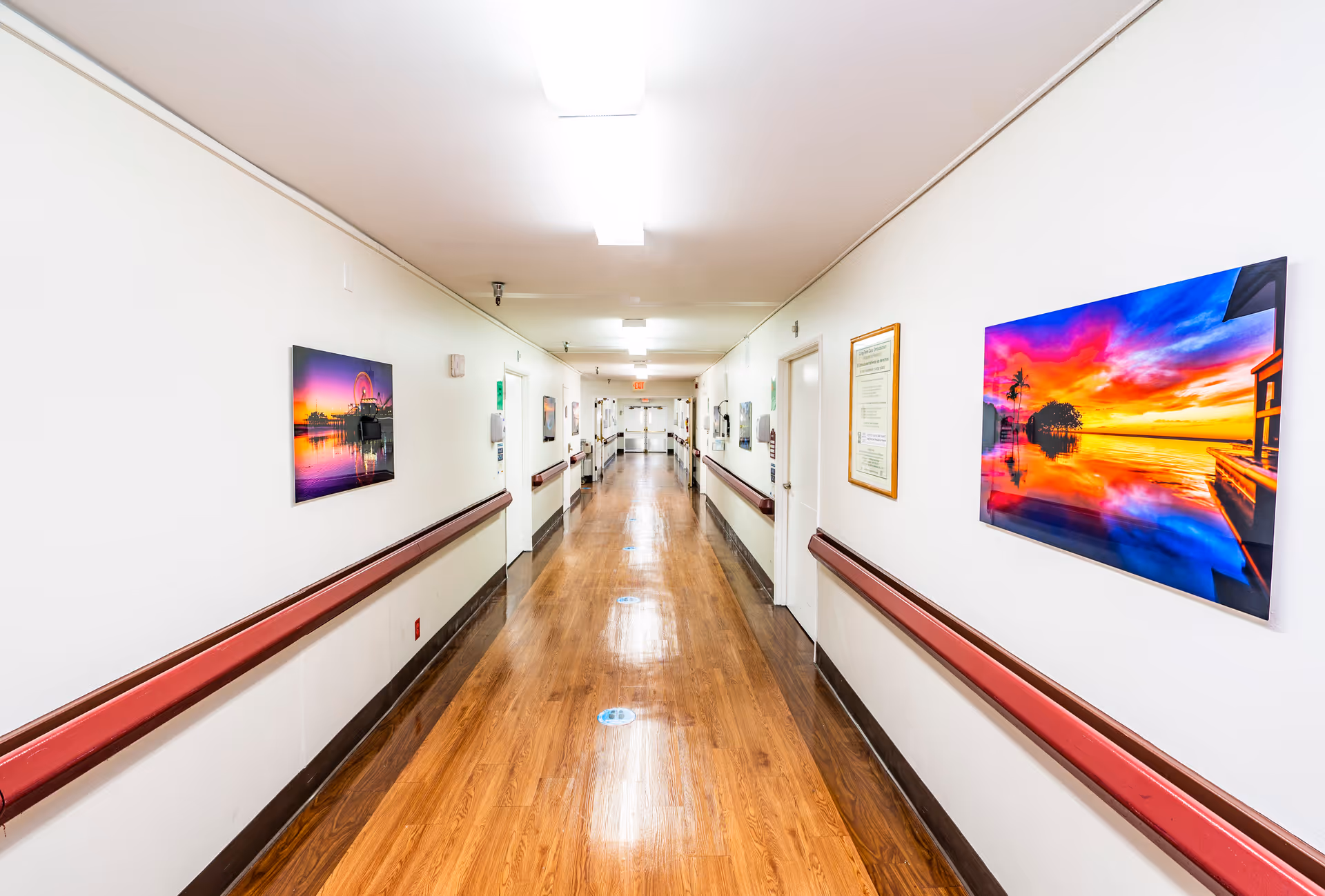 Well-lit long interior hallway in a care facility with wood floors, handrails and colorful artwork on the walls.
