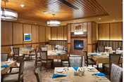 A dining room with multiple round tables covered with beige tablecloths, each set with plates, glasses, and blue napkins. The room features wooden paneling on the walls, a fireplace with a framed picture above it, and two ceiling light fixtures. Chairs with patterned upholstery surround the tables.