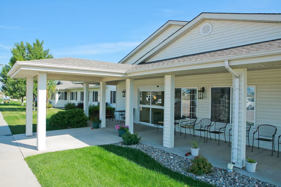 Exterior view of a senior living facility with a covered porch area featuring several black metal chairs and potted plants. The building has white siding, a sloped roof, and large windows. There is a well-maintained lawn and a sidewalk leading to the entrance.