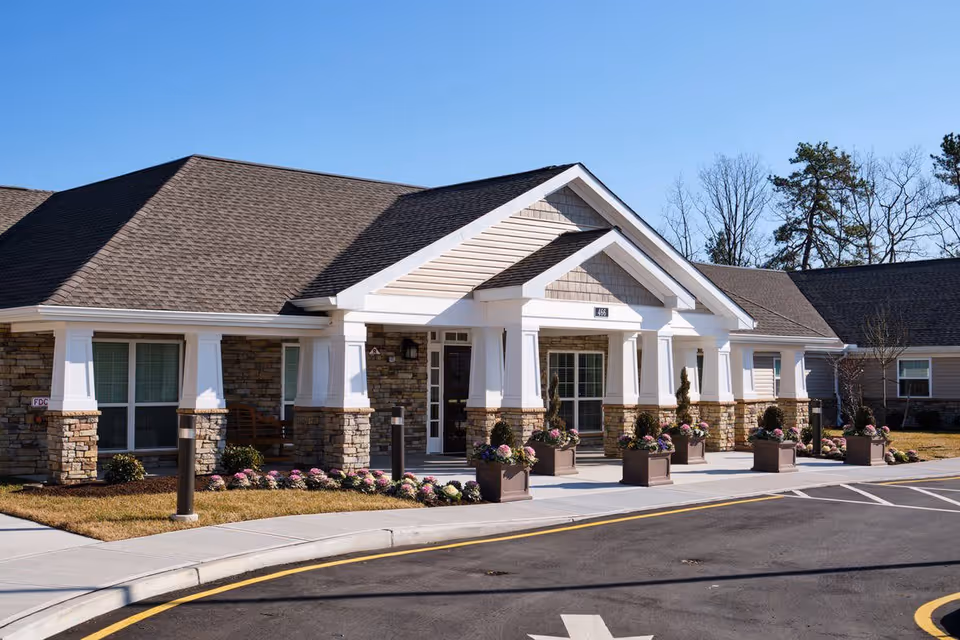 Exterior front view of a single-story senior living facility building with stone and beige siding, white columns, and a covered entrance. There are flower pots with pink and purple flowers lining the walkway and a clear blue sky in the background.