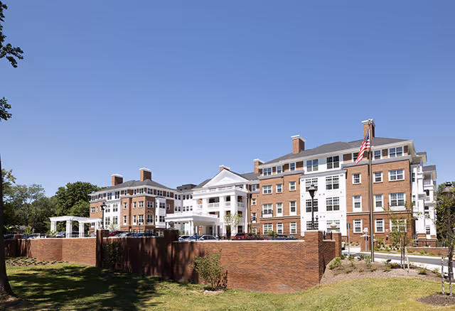 Exterior view of a large senior living facility building with red brick and white accents, multiple windows, and a prominent entrance with columns. There is an American flag on a flagpole in front of the building, and a well-maintained lawn and trees surrounding the property under a clear blue sky.