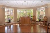 Small chapel interior with a wooden altar topped by a cross, flanked by chairs and large bay windows.