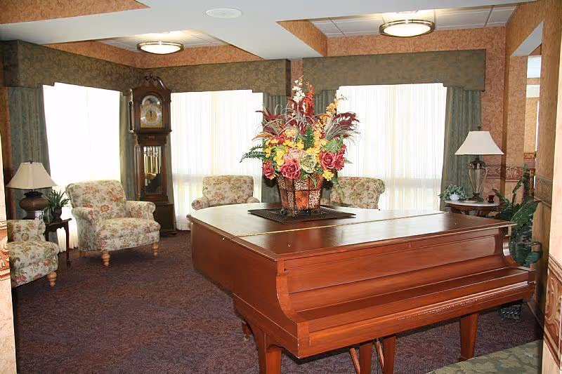 A cozy living room area with a large wooden grand piano in the center topped with a floral arrangement. Surrounding the piano are floral upholstered armchairs, a tall grandfather clock, side tables with lamps, and large windows with curtains allowing natural light to fill the room.
