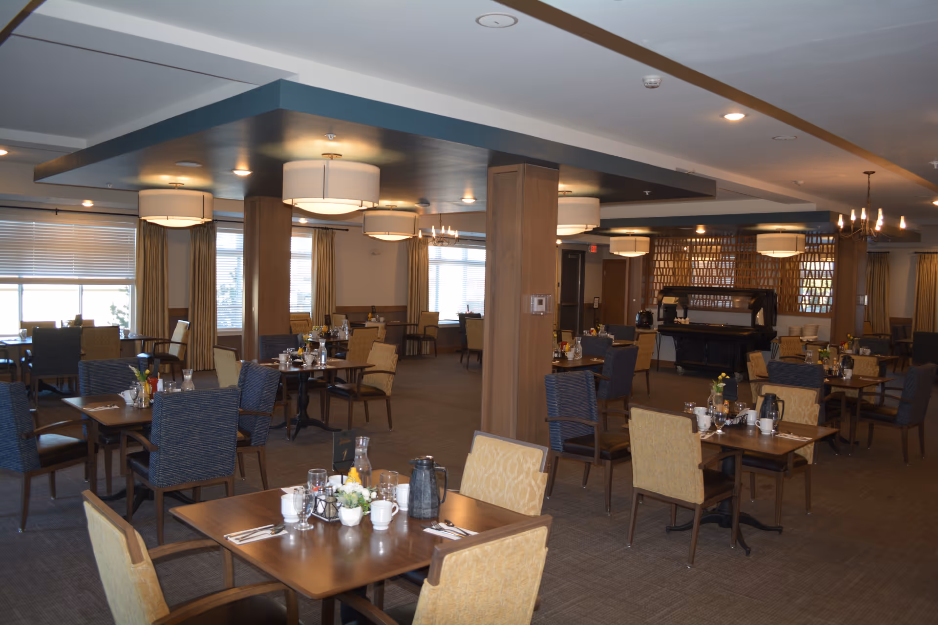 A spacious dining room in a senior living facility with multiple wooden tables and chairs arranged neatly. The tables are set with cups, glasses, utensils, and small flower arrangements. The room has large windows with curtains, modern ceiling lights, and wooden pillars. The atmosphere is warm and inviting.
