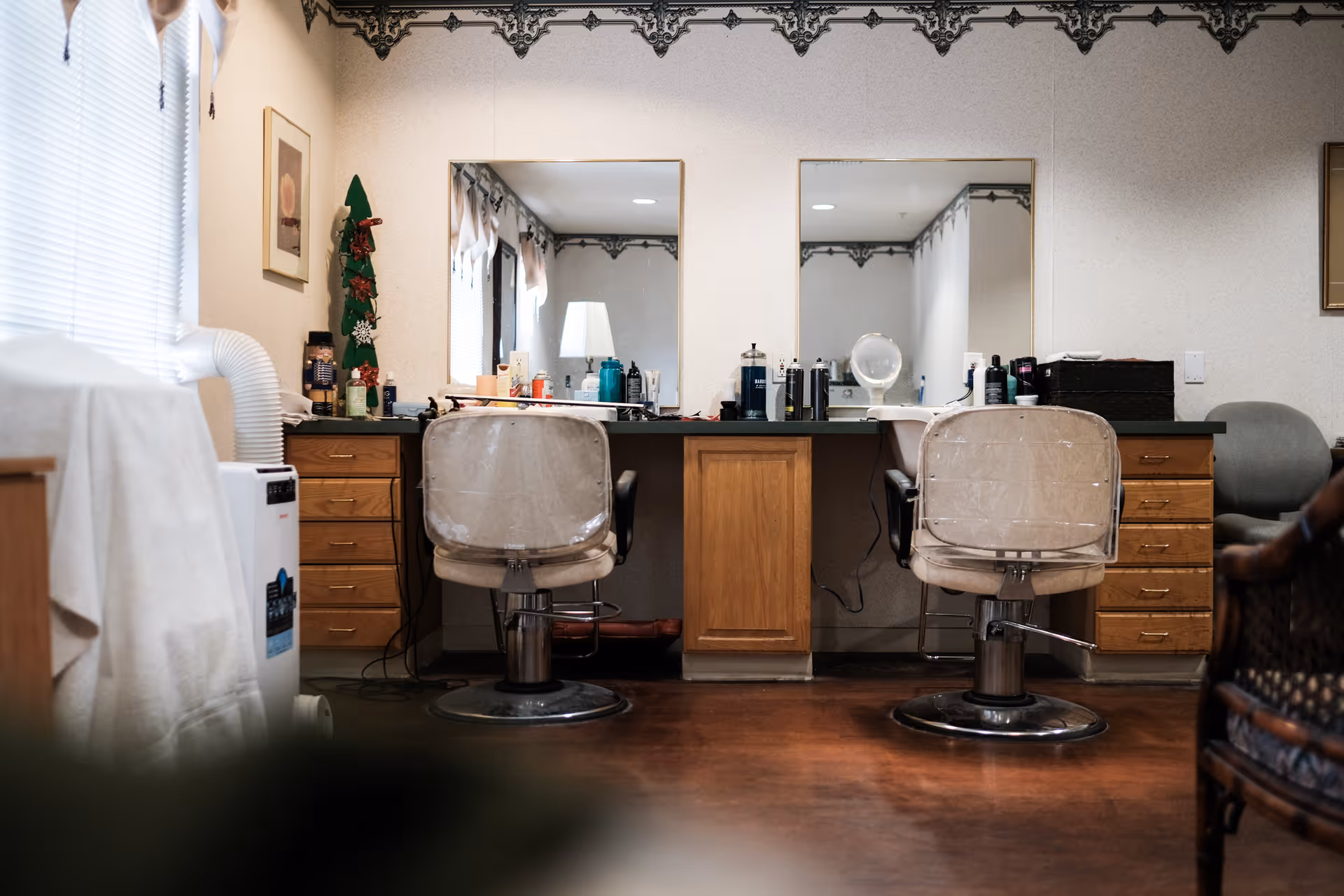 Interior view of a salon area with two salon chairs facing a counter with mirrors. The counter has various hair and beauty products, and there are wooden drawers underneath. The room has a decorative border near the ceiling and a window with blinds on the left side.