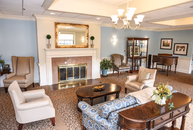 A cozy living room with a fireplace centered on the far wall, flanked by two small potted plants. The room features a patterned blue sofa, beige armchairs, a wooden coffee table, and a wooden side table with a flower arrangement. There is a chandelier overhead, a glass display cabinet, framed pictures on the walls, and a grand piano in the background.