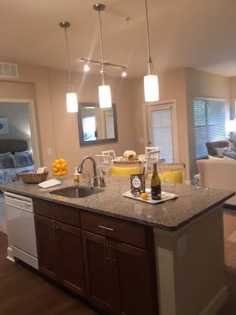 Interior view of a senior living facility kitchen area with a granite countertop island featuring a sink, a bottle of wine, and a tray with glasses. Three pendant lights hang above the island. In the background, there is a dining area with chairs and a table, a living room with seating, and a bedroom visible through an open doorway.