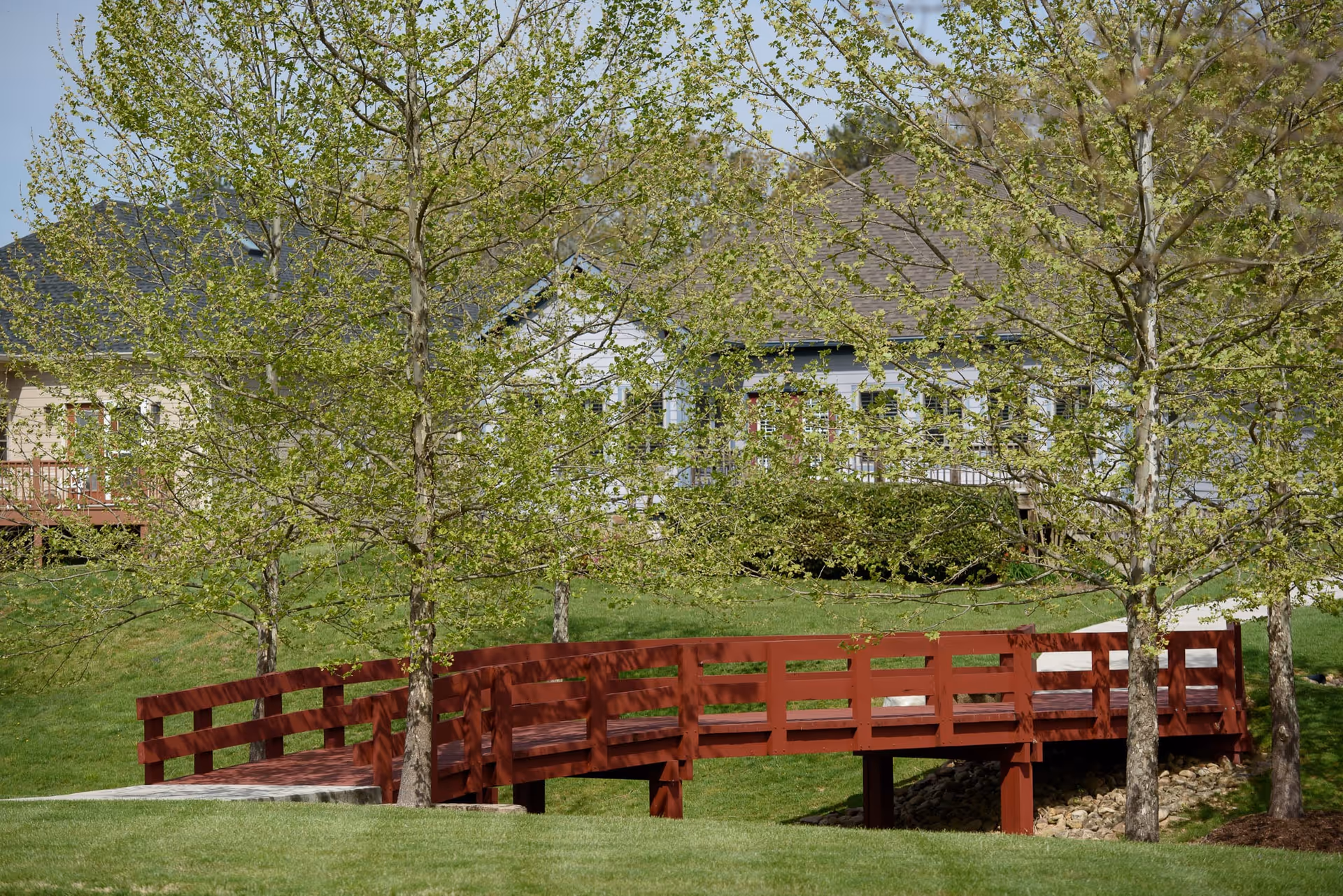 Red wooden footbridge spanning a small landscaped area with trees and a residential building in the background.