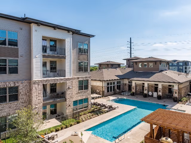 Outdoor view of a senior living facility featuring a large rectangular swimming pool surrounded by lounge chairs and a pergola. The pool area is adjacent to a multi-story residential building with balconies and a single-story building with a tiled roof. The sky is clear and blue.