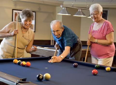 Three elderly individuals playing pool together in a recreational room, smiling and enjoying the game.