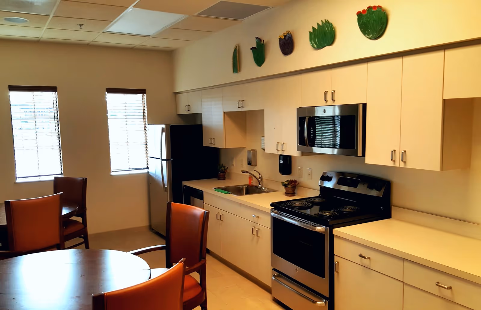 A kitchen area with white cabinets, a stainless steel refrigerator, stove, and microwave. There is a sink with a faucet and small potted plants on the countertop. In the foreground, there are round wooden tables with orange cushioned chairs. Two windows with blinds are visible on the left wall, and decorative green and purple wall hangings are above the cabinets.